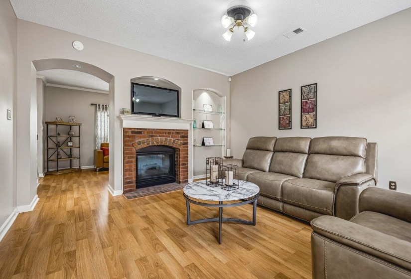 Living area featuring wood-finish flooring, a brick fireplace with a white mantel, an arched entryway, and a built-in recessed shelf with glass shelving