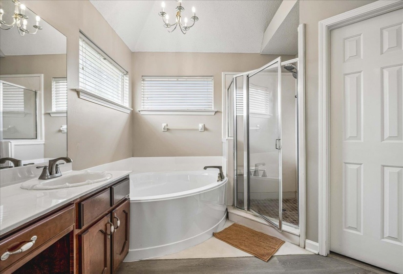 Bathroom featuring a large corner soaking tub, separate glass-enclosed shower with tile flooring, wood-finish vanity with an integrated sink and white countertop, and dual windows with blinds