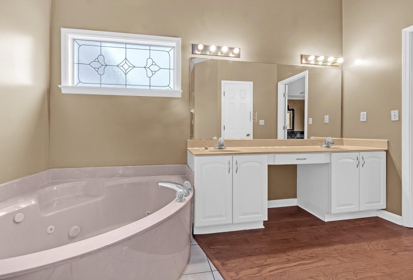 Bathroom featuring a built-in corner soaking tub, a decorative leaded glass window, dual vanities with white cabinetry, a large wall-mounted mirror, and wood-finish flooring