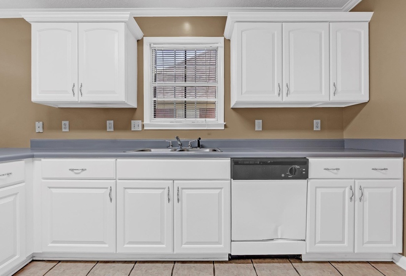 Kitchen featuring white cabinetry with silver hardware, grey countertops, a stainless steel sink with a chrome faucet, and a built-in white dishwasher with a black control panel