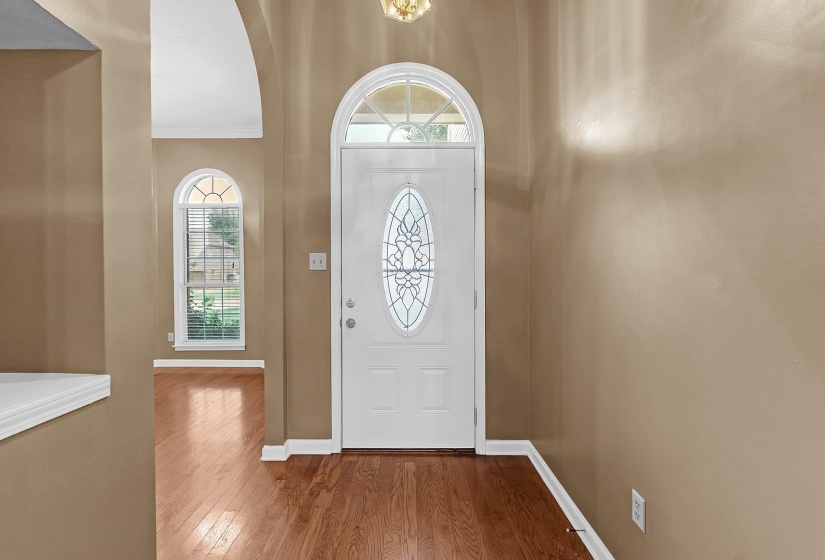 Hardwood-finish flooring extending from the entryway into other spaces, featuring a white door with decorative glass inlay, and an arched transom window