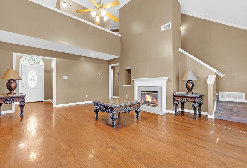 Spacious living area featuring high ceilings, wood-finish flooring, a fireplace with white mantel, a decorative front door with an oval glass insert, and an upper-level balcony overlooking the space